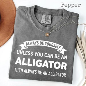 May include: A grey t-shirt with the slogan "ALWAYS BE YOURSELF UNLESS YOU CAN BE AN ALLIGATOR THEN ALWAYS BE AN ALLIGATOR" in white. The shirt has a crew neck and short sleeves. A silver necklace with a pendant is on the shirt.