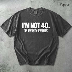 Könnte beinhalten: Pfefferfarbenes T-Shirt mit dem weißen Text "I'M NOT 40. I'M TWENTY-TWENTY." in Großbuchstaben. Das Shirt hat einen Rundhalsausschnitt und kurze Ärmel. Der Stoff scheint vorgewaschen und leicht verblichen zu sein.