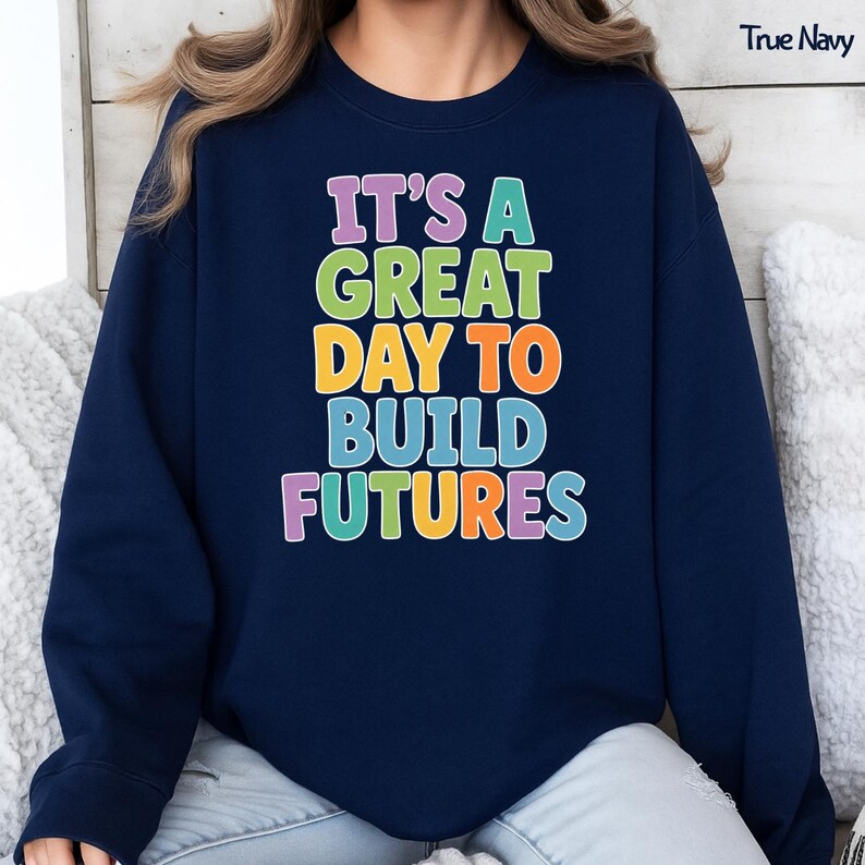 Puede incluir: Sudadera azul marino con el texto "IT'S A GREAT DAY TO BUILD FUTURES" en letras coloridas y de burbujas. El texto est&aacute; apilado verticalmente, cada palabra en un color diferente. Un estilo c&oacute;modo e informal.