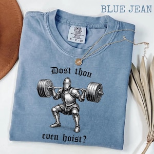 May include: A blue jean colored t-shirt with the text "Dost thou even hoist?" and a graphic of a knight lifting weights. The shirt has a crew neck and short sleeves. A gold necklace with a pendant is draped across the shirt.
