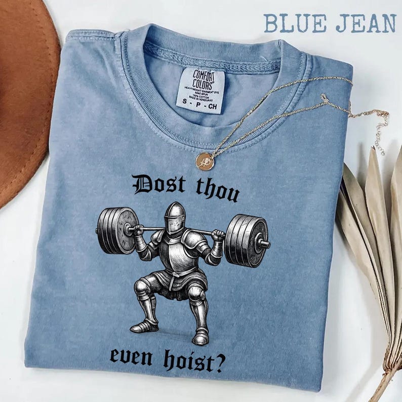 May include: A blue jean colored t-shirt with the text "Dost thou even hoist?" and a graphic of a knight lifting weights. The shirt has a crew neck and short sleeves. A gold necklace with a pendant is draped across the shirt.