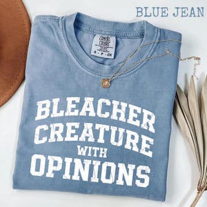 Könnte beinhalten: Ein blaues Jeans-T-Shirt mit dem Text "BLEACHER CREATURE WITH OPINIONS" in weißen Blockbuchstaben. Eine goldene Halskette mit Anhänger liegt über dem Shirt. Das Shirt ist aus weichem Material.