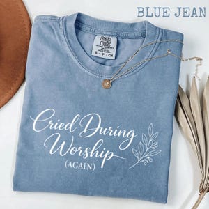 Puede incluir: Una camiseta azul vaquero con el texto "Cried During Worship (Again)" en escritura blanca. Un collar dorado con un colgante descansa sobre la camiseta. La camiseta está doblada y tiene una etiqueta Comfort Colors.