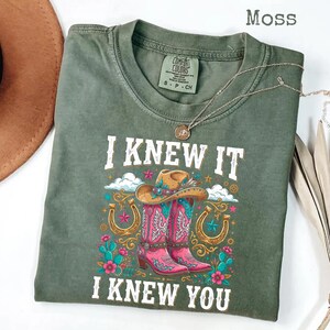 May include: A moss green short-sleeve t-shirt featuring a graphic print of a cowboy hat, pink western boots, horseshoes, and cacti. The text reads I KNEW IT I KNEW YOU. The shirt has a crew neckline and a Comfort Colors brand label at the collar.