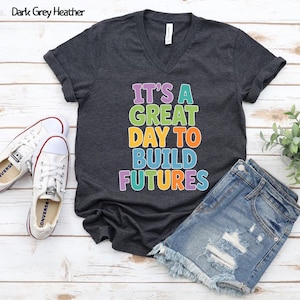 Puede incluir: Camiseta de cuello en V gris oscuro jaspeado con el texto multicolor "IT'S A GREAT DAY TO BUILD FUTURES". Se combina con pantalones cortos vaqueros y zapatillas de lona blancas.