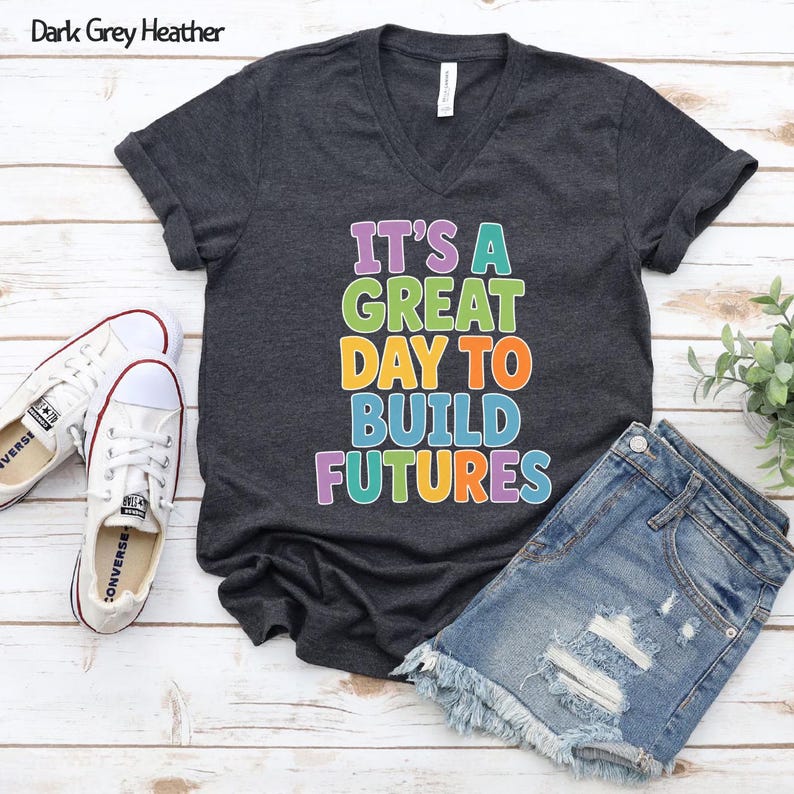 Puede incluir: Camiseta de cuello en V gris oscuro jaspeado con el texto multicolor "IT'S A GREAT DAY TO BUILD FUTURES". Se combina con pantalones cortos vaqueros y zapatillas de lona blancas.