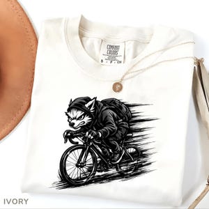 Pode incluir: Camiseta de cor marfim com uma ilustração a preto e branco de um lobo a andar de bicicleta. O lobo está a usar um casaco com capuz e uma mochila, com linhas de movimento atrás da bicicleta. Um colar de ouro e um chapéu são visíveis.