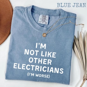 以下が含まれることがあります： 「I'M NOT LIKE OTHER ELECTRICIANS (I'M WORSE)」の文字が入った、ブルーデニムのComfort Colors Tシャツ。ペンダント付きのゴールドネックレスがシャツの上にかけられています。シャツのサイズはスモールです。