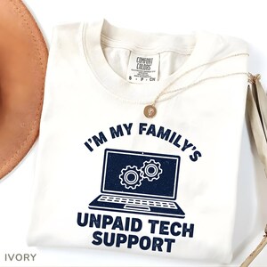 May include: Ivory t-shirt with navy blue text: "I'M MY FAMILY'S UNPAID TECH SUPPORT". The design features a laptop graphic with two gears. A gold necklace with a pendant is draped across the shirt. The shirt is a Comfort Colors brand.