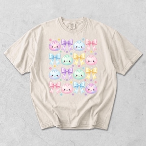 May include: A beige short-sleeve t-shirt featuring a repeating pattern of pastel-colored cartoon dumplings with smiling faces, interspersed with various bows in shades of pink, blue, yellow, and purple, along with small colorful flower accents.