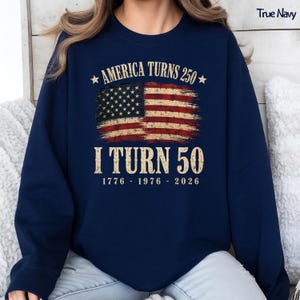 May include: Navy blue sweatshirt with the text "America Turns 250" and "I Turn 50" in gold lettering. The design includes a distressed American flag graphic. The years 1776, 1976, and 2026 are printed below the text.