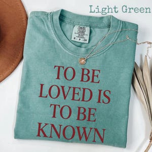 May include: Light green t-shirt with the text "TO BE LOVED IS TO BE KNOWN" in red. The shirt has a crew neck and short sleeves. A silver necklace with a small pendant is draped across the shirt.