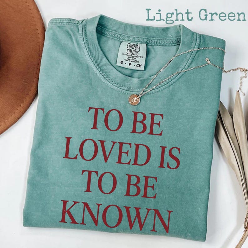 May include: Light green t-shirt with the text "TO BE LOVED IS TO BE KNOWN" in red. The shirt has a crew neck and short sleeves. A silver necklace with a small pendant is draped across the shirt.
