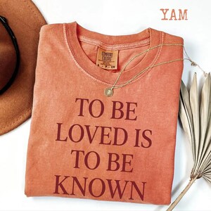 May include: A coral-colored t-shirt with the text "TO BE LOVED IS TO BE KNOWN" in a darker shade. A gold necklace with a pendant rests on the shirt. The shirt is from Comfort Colors.