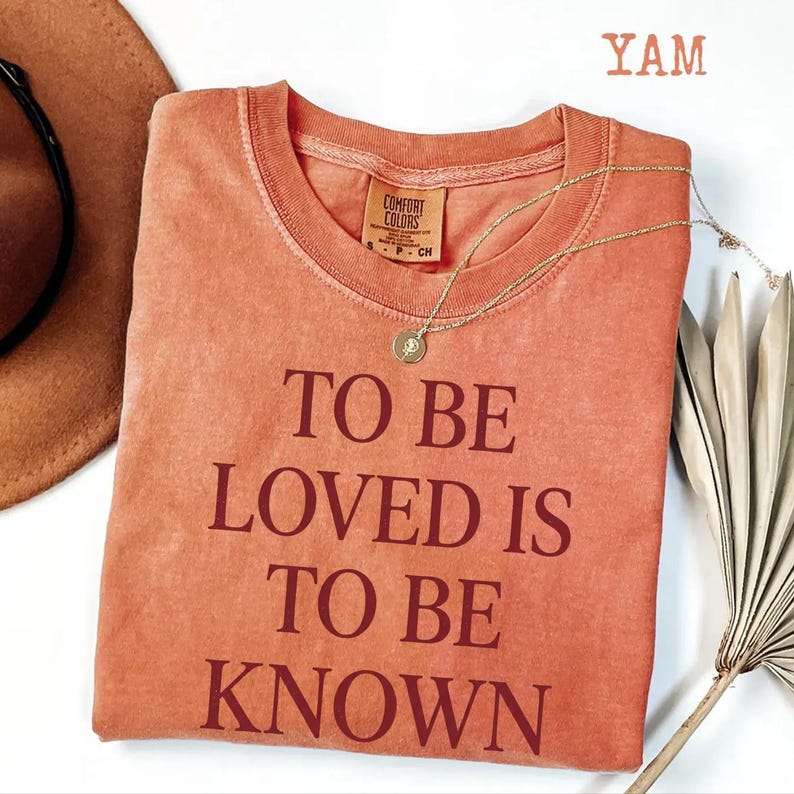 May include: A coral-colored t-shirt with the text "TO BE LOVED IS TO BE KNOWN" in a darker shade. A gold necklace with a pendant rests on the shirt. The shirt is from Comfort Colors.