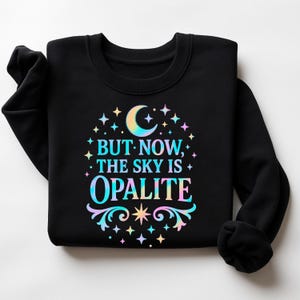 Pode incluir: Camisola preta de gola redonda com um gráfico colorido. O gráfico apresenta uma lua crescente, estrelas e o texto "BUT NOW, THE SKY IS OPALITE" num gradiente arco-íris. O design tem um tema celestial.