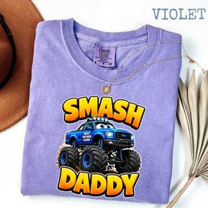 Może przedstawiać: Fioletowa koszulka z niebieską grafiką monster trucka i napisem "SMASH DADDY" w kolorze ż&oacute;łtym i pomarańczowym. Na ciężar&oacute;wce widnieje napis "BEST FATHER EVER". Złoty naszyjnik spoczywa na koszulce.