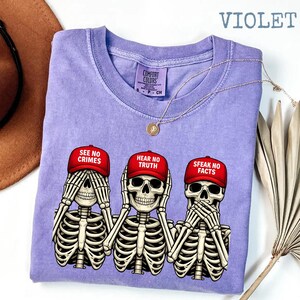 May include: Violet-colored t-shirt featuring three skeleton figures in the 'see no evil, hear no evil, speak no evil' pose. Each skeleton wears a red baseball cap with a different phrase: 'See No Crimes,' 'Hear No Truth,' and 'Speak No Facts.' A gold necklace is draped across the shirt.