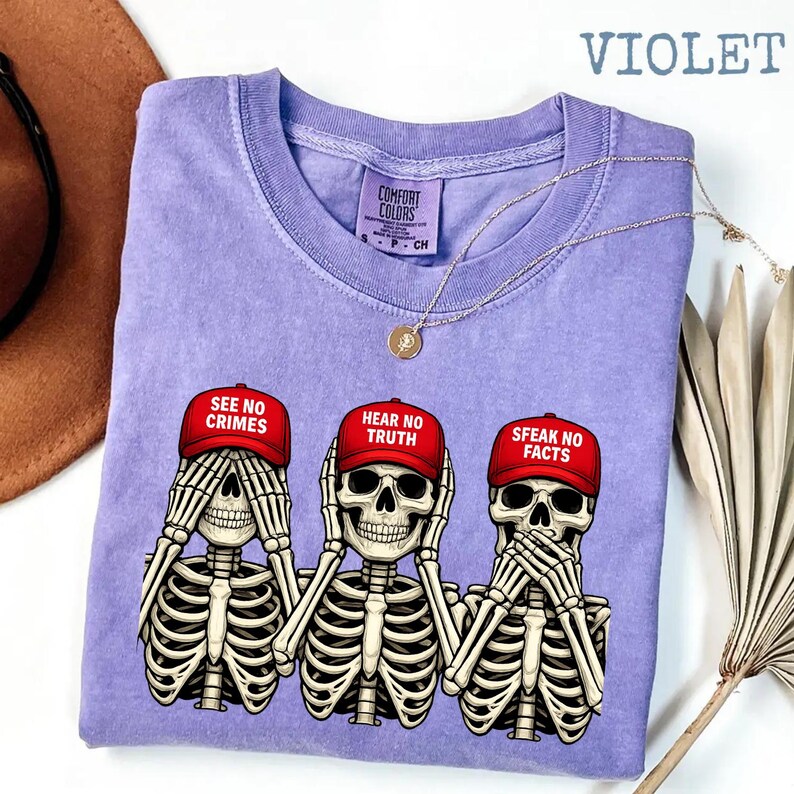 May include: Violet-colored t-shirt featuring three skeleton figures in the 'see no evil, hear no evil, speak no evil' pose. Each skeleton wears a red baseball cap with a different phrase: 'See No Crimes,' 'Hear No Truth,' and 'Speak No Facts.' A gold necklace is draped across the shirt.