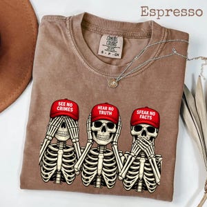 May include: Espresso-colored t-shirt featuring three skeleton figures in the 'see no evil, hear no evil, speak no evil' pose. Each skeleton wears a red baseball cap with a different phrase: 'SEE NO CRIMES,' 'HEAR NO TRUTH,' and 'SFEAK NO FACTS.'