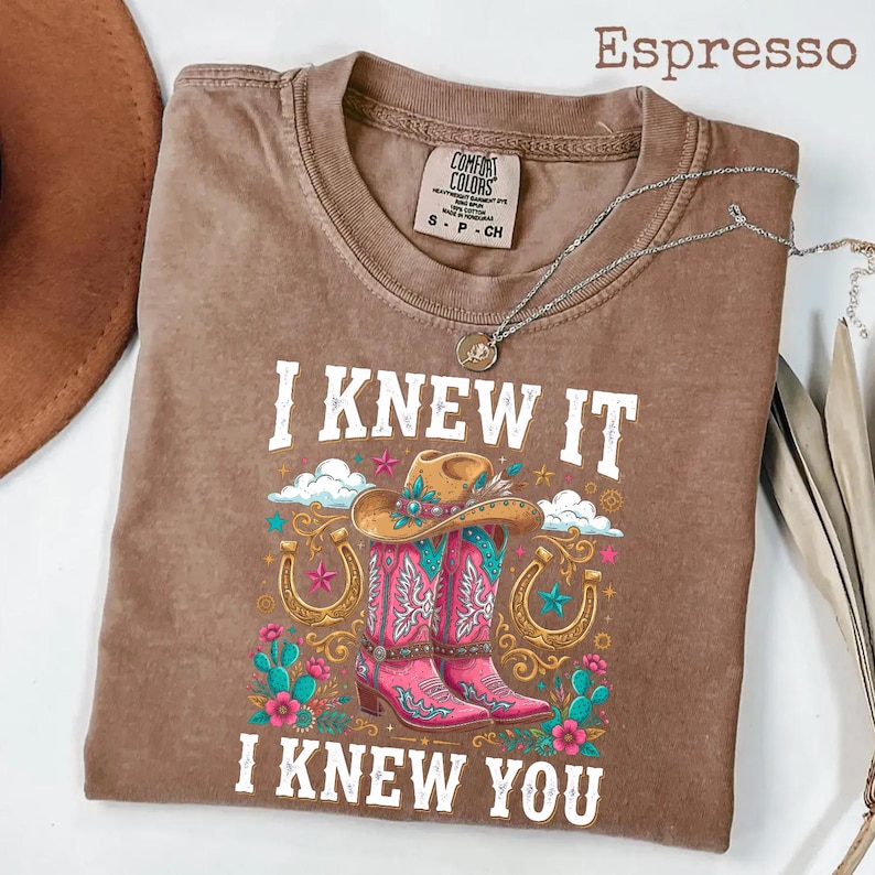 May include: An espresso-colored short-sleeve t-shirt featuring a graphic print of pink cowboy boots, a tan cowboy hat, horseshoes, and cacti. The text "I KNEW IT" and "I KNEW YOU" is printed in white above and below the colorful western-themed illustration.
