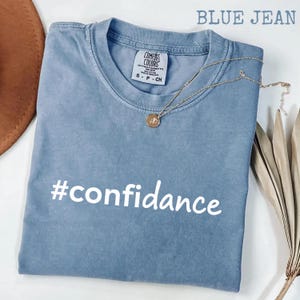 Peut inclure: Un t-shirt bleu jean Comfort Colors avec le mot "#confidance" en blanc. Un collier en or avec un pendentif est drapé sur le t-shirt. Le t-shirt est plié proprement sur une surface blanche.