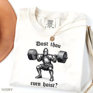 May include: Ivory-colored t-shirt with the text "Dost thou even hoist?" and a graphic of a knight lifting weights. The shirt has a Comfort Colors tag and a gold necklace. The shirt is a size small.