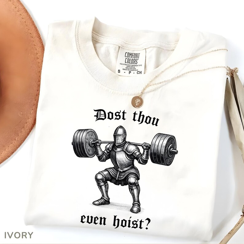 May include: Ivory-colored t-shirt with the text "Dost thou even hoist?" and a graphic of a knight lifting weights. The shirt has a Comfort Colors tag and a gold necklace. The shirt is a size small.