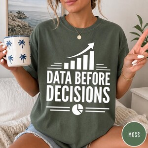 K&ouml;nnte beinhalten: Ein moosgr&uuml;nes T-Shirt mit Rundhalsausschnitt und einem wei&szlig;en Grafikdruck, der ein Balkendiagramm mit einem Pfeil nach oben, den Text "DATA BEFORE DECISIONS" und ein Tortendiagramm-Symbol zeigt, kombiniert mit einer wei&szlig;en Tasse mit blauem Palmenmuster.