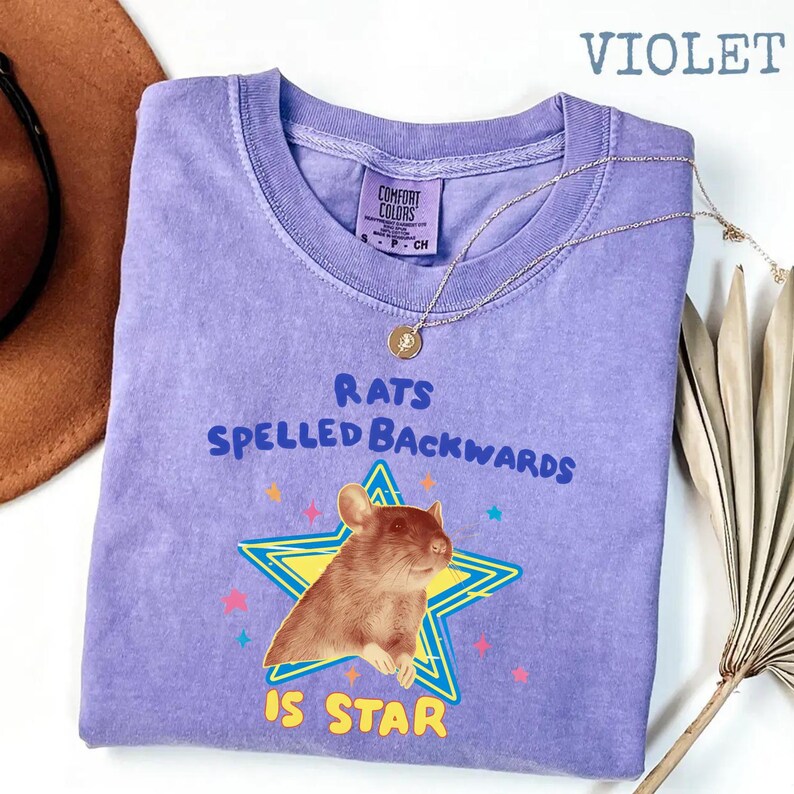 May include: A violet-colored t-shirt with the text "RATS SPELLED BACKWARDS IS STAR" and a graphic of a rat inside a star. The shirt also features a Comfort Colors tag and a gold necklace.
