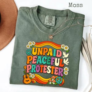 以下が含まれることがあります： モスグリーンのTシャツ。カラフルなレトロスタイルの文字で「UNPAID PEACEFUL PROTESTER」と書かれています。デザインには、平和のシンボル、ギター、花、キノコ、ハトが含まれています。ペンダント付きのシルバーネックレスがシャツの上にかけられています。