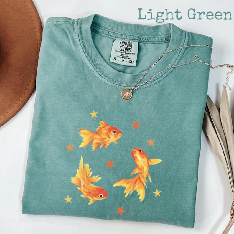 May include: Light green Comfort Colors t-shirt featuring three orange goldfish and orange stars. The shirt has a crew neck and short sleeves. A silver necklace with a pendant is draped across the shirt.