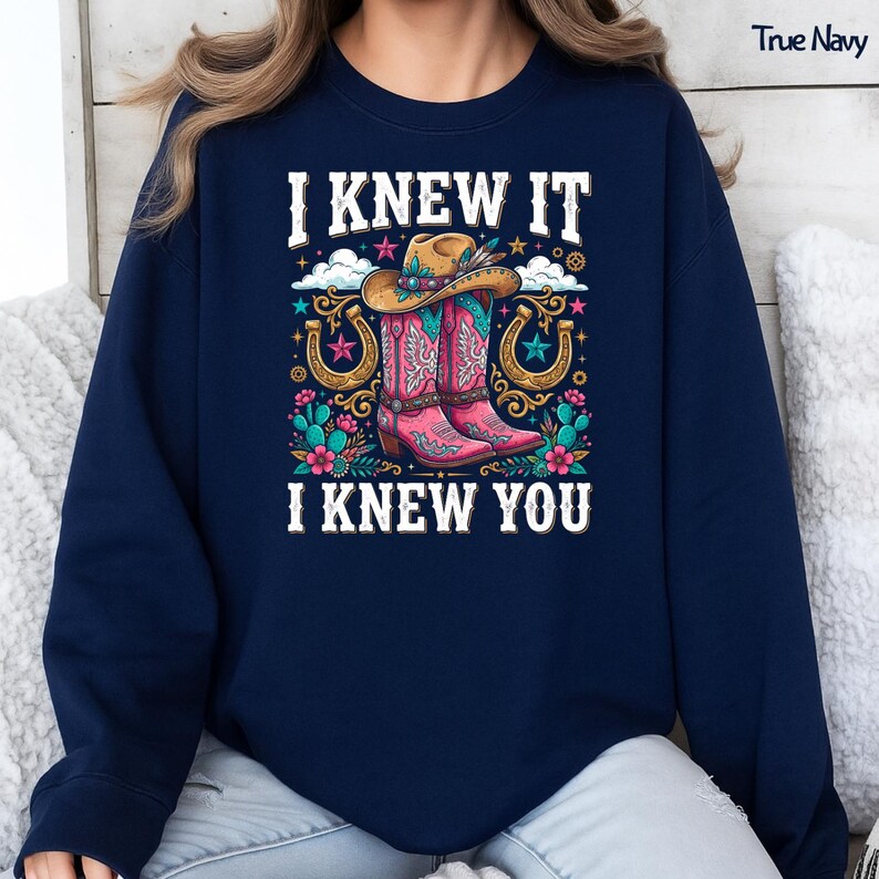 May include: A true navy long-sleeve sweatshirt featuring a graphic print with the text I KNEW IT I KNEW YOU. The design includes a tan cowboy hat, a pair of pink cowboy boots, two gold horseshoes, teal cacti, and colorful flowers and stars.