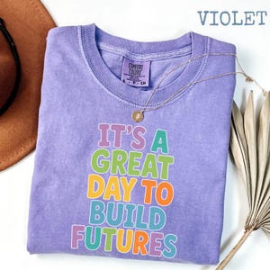 Puede incluir: Una camiseta violeta Comfort Colors con el texto "IT'S A GREAT DAY TO BUILD FUTURES" en letras de colores. Un collar dorado con un colgante descansa sobre la camiseta. La palabra "VIOLET" est&aacute; en la esquina superior derecha.
