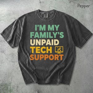 Puede incluir: Camiseta color pimienta con el texto "I'M MY FAMILY'S UNPAID TECH SUPPORT" en verde azulado, blanco, amarillo y naranja. El diseño incluye un icono de monitor de ordenador con una llave inglesa y un destornillador.