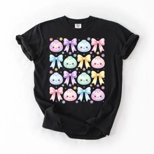 May include: A black t-shirt featuring a repeating pattern of pastel-colored, smiling dumpling characters and bows in shades of pink, blue, purple, and yellow, accented with small colorful flower icons arranged in a grid across the chest.