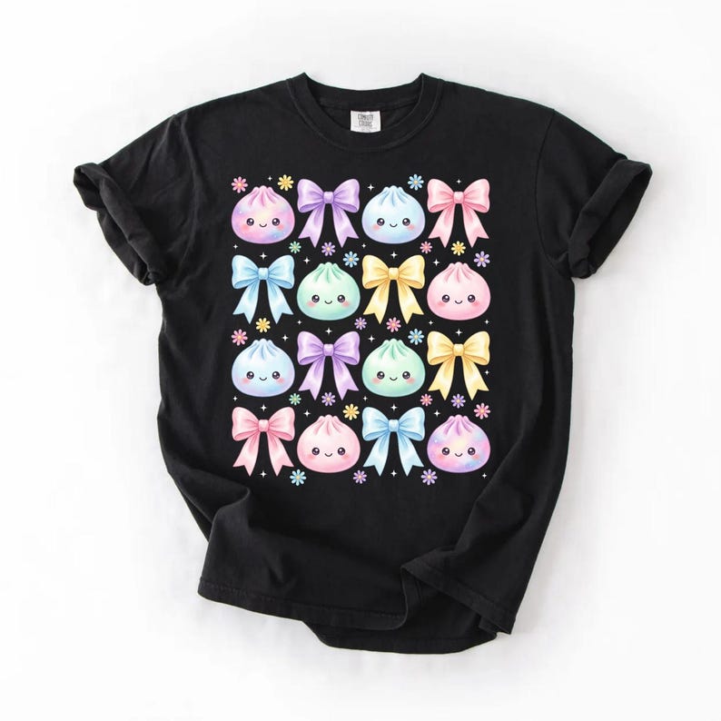 May include: A black t-shirt featuring a repeating pattern of pastel-colored, smiling dumpling characters and bows in shades of pink, blue, purple, and yellow, accented with small colorful flower icons arranged in a grid across the chest.