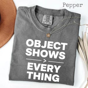 May include: A gray t-shirt with the text "OBJECT SHOWS EVERYTHING" in white block letters. The shirt has a crew neck and short sleeves. A silver necklace with a pendant is on the shirt. The shirt is folded on a white surface.