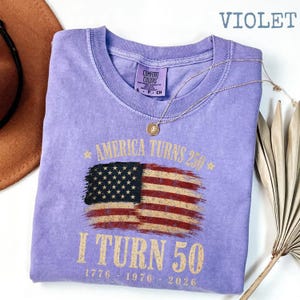 May include: Violet Comfort Colors t-shirt with a distressed American flag graphic. The shirt reads "America Turns 250, I Turn 50" with the years 1776, 1976, and 2026. A gold necklace and a brown hat are also in the image.