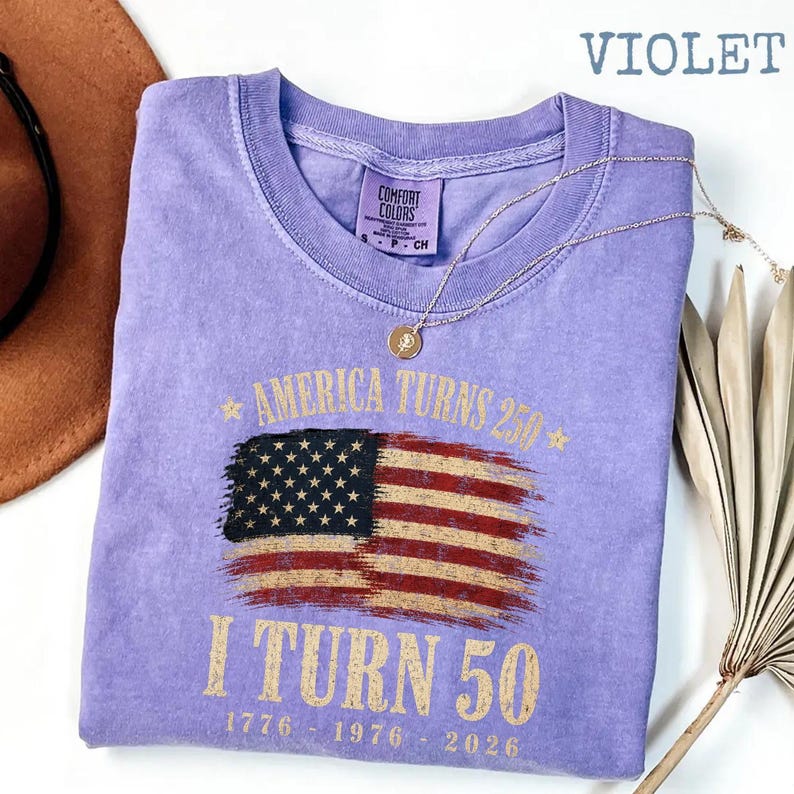 May include: Violet Comfort Colors t-shirt with a distressed American flag graphic. The shirt reads "America Turns 250, I Turn 50" with the years 1776, 1976, and 2026. A gold necklace and a brown hat are also in the image.