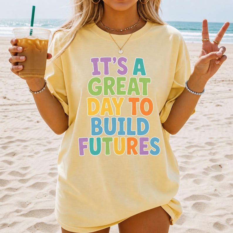 Puede incluir: Camiseta amarillo claro con el texto "IT'S A GREAT DAY TO BUILD FUTURES" en letras de colores. La camiseta se lleva en un entorno de playa.