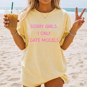 May include: A light yellow t-shirt with the text "SORRY GIRLS I ONLY DATE MODELS" in pink. The shirt is worn on a beach with a drink in one hand and a peace sign made with the other hand.