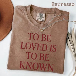 May include: Espresso-colored t-shirt with the text "TO BE LOVED IS TO BE KNOWN" in red. The shirt has a crew neck and short sleeves. A silver necklace with a pendant is draped across the shirt.