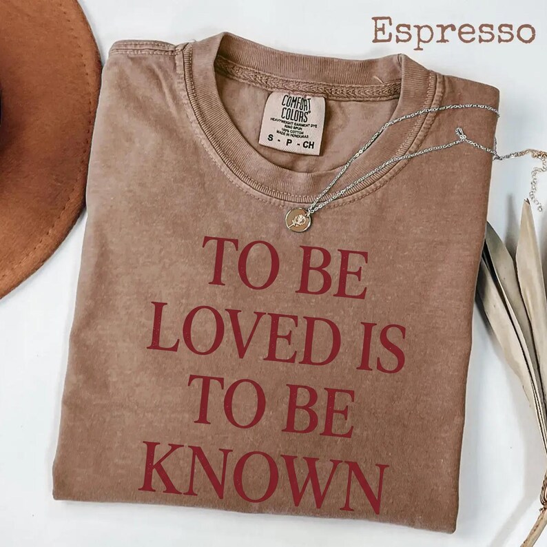 May include: Espresso-colored t-shirt with the text "TO BE LOVED IS TO BE KNOWN" in red. The shirt has a crew neck and short sleeves. A silver necklace with a pendant is draped across the shirt.
