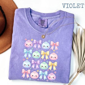 May include: A violet short-sleeved t-shirt featuring a graphic print of various pastel-colored bows and smiling dumpling characters. The shirt has a crew neckline and a Comfort Colors label. The text VIOLET is printed in the upper right corner.