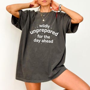 Könnte beinhalten: Ein anthrazitfarbenes Oversize-T-Shirt mit dem weißen Schriftzug "wildly unprepared for the day ahead" auf der Brust, akzentuiert durch kleine Sternmotive. Das Kleidungsstück hat kurze Ärmel, einen Rundhalsausschnitt und eine lockere Passform.
