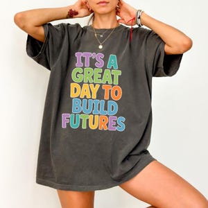 Puede incluir: Camiseta gris oscuro con el texto "IT'S A GREAT DAY TO BUILD FUTURES" en letras de colores. La camiseta tiene un corte holgado y mangas cortas. El texto es en morado, verde, naranja, azul y amarillo.