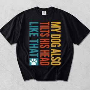 May include: A black short-sleeve t-shirt featuring vertical text in yellow, orange, and teal that reads MY DOG ALSO TILTS HIS HEAD LIKE THAT, accompanied by a white paw print graphic positioned below the final word of the text.
