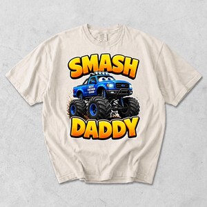 Może przedstawiać: Kremowa koszulka z niebieską grafiką monster trucka i napisem "SMASH DADDY" w kolorze ż&oacute;łtym i pomarańczowym. Samoch&oacute;d ma napis "BEST FATHER EVER" z boku.
