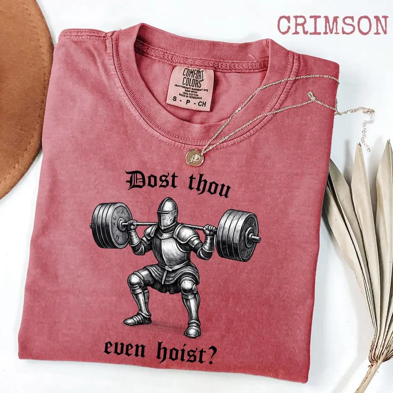 May include: Crimson-colored t-shirt with a graphic of a knight lifting weights, with the text "Dost thou even hoist?" The shirt has a crew neck and short sleeves. A gold necklace with a pendant is draped across the shirt.